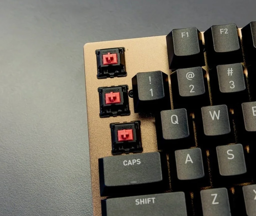 Mechanical keyboard close-up: black keycaps on a bronze metal plate with three keys removed, showing red linear switches, on a dark grey desk