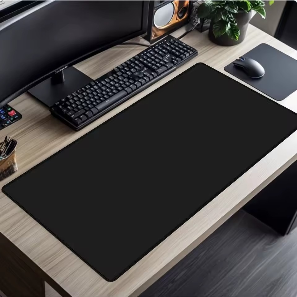Large black extended desk mat on a light wood desk with keyboard, monitor, and mouse setup